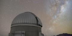 The Milky Way and night sky above Rubin Observatory's Auxiliary Telescope. The auxiliary telescope is at the lower left, and the night sky is sprinkled with stars. The denser area of the Milky Way extends upward on the right, bulging wider toward the top. Dark tendrils of dust clouds weave through the bulge.